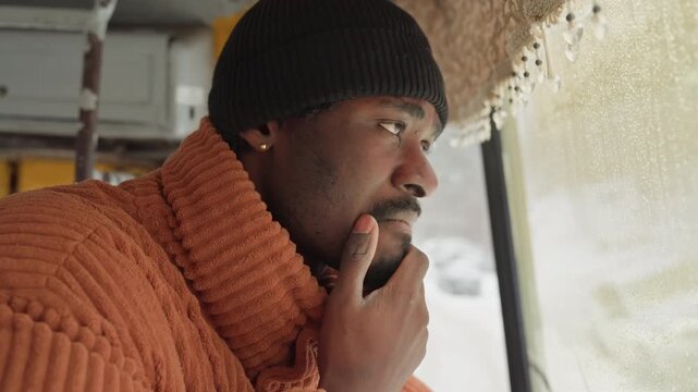 Black man wiping eyes on bus, orange sweater and black beanie, frosted glass with raindrops, hand to face, closeup profile showing moment of vulnerability, interior light and winter commute tension