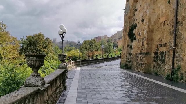 Scenic view of a winding street with stone wall and lampposts, showcasing lush greenery and distant Lombardy Castle in Enna, Sicily, under a cloudy sky