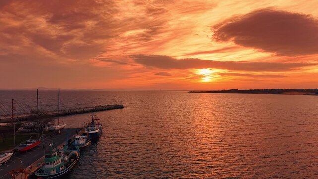 Moving over the waterscape colored orange at sunset. Cloudy orange sky in the backdrop. Aerial view on marina with boats.
