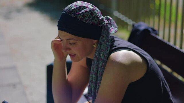 Headscarf woman in chemotherapy sitting in park talking on phone video, animated expression and hand movement, real candid moment of connection in warm sunlight