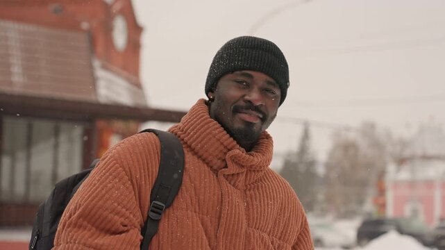 Black man in orange sweater adjusts glasses on snowy city sidewalk, removes glasses, points toward camera and smiles with backpack and beanie portrays student, commuter, traveler and friend amid soft