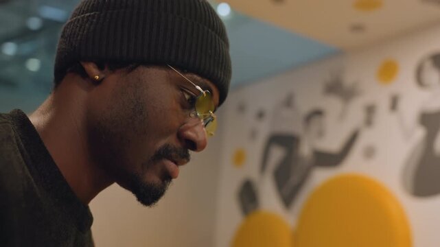 Man adjusts glasses by coat rack, closeup portrait with knit beanie, subtle smile and thoughtful gaze, tinted frames and stud earring, warm bokeh mural background and contemplative mood