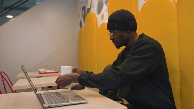 Black man coding on laptop cafe focused developer debugging code with coffee cup on table, yellow acoustic wall, glasses and beanie, startup energy, collaborative workspace atmosphere