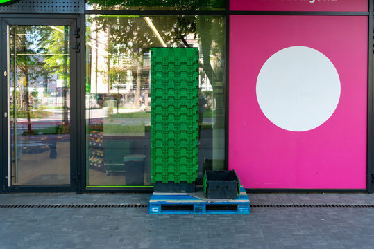 Stacked green plastic produce crates stand on a pallet outside a glass grocery storefront with a bright pink wall panel and large white circle. Useful for retail supply, food store logistics and urban