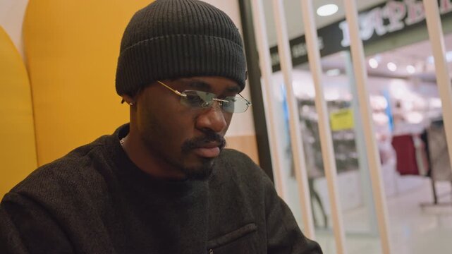 Black man in beanie working laptop, seated in yellow booth at mall lounge with warm ambient lighting, reading documents and typing notes, calm focused energy of freelance writer or student, glasses