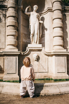Toddler child looking at classical fountain statue in historic park on sunny day