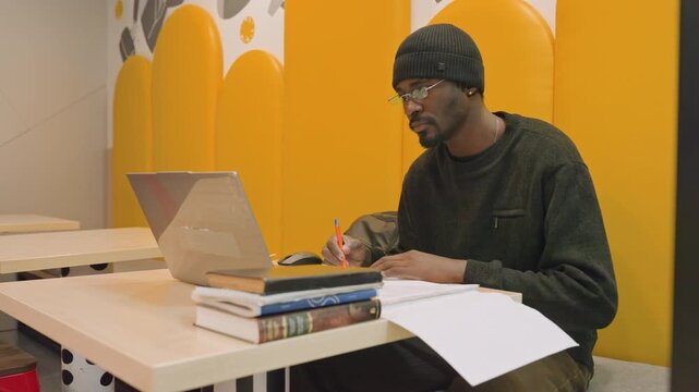 Young man studying at laptop in yellow study booth, flipping pages and writing notes, juggling textbooks while preparing for exam, sketching ideas for freelance design project, researching references