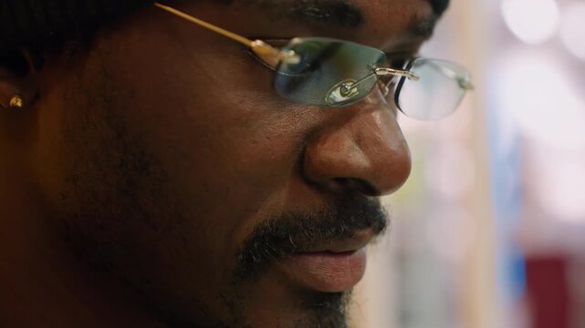 Closeup man wearing glasses focused on sketches, yellowlit studio, gold frames glinting, beanie, serious gaze, careful hands, designer at work, cinematic shallow depth, textured grain, urban craft