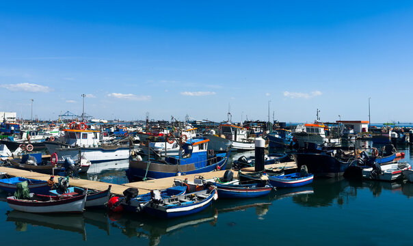 Colorful traditional fishing boats moored in the calm, reflective water of Setubal harbor, Portugal. Stone steps and a beached boat on the shore. Setubal, Portugal. 