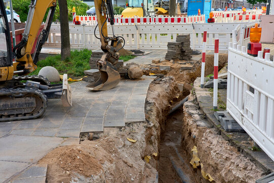 Excavator working on a narrow trench for utility pipelines on a city sidewalk. Road construction site with safety barriers and paving stones.