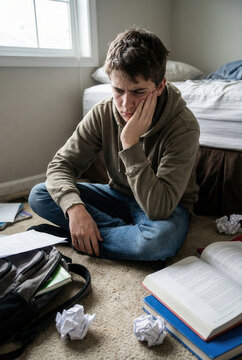 Teenager boy sits on the floor, looking distressed. Overwhelmed student guy surrounded by open textbooks, crumpled papers, and a backpack, feels intense academic pressure and mental fatigue