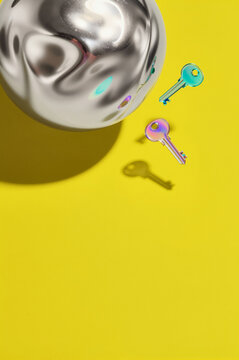 Iridescent keys cast sharp shadows onto a saturated mustard yellow plane next to a large, molten silver orb with fluid, mercury-like undulations. High-contrast lighting creates deep silhouettes and