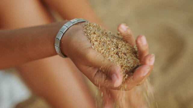 Cupped hand holding warm beach sand, silver bracelet glint, sunlit grains slipping through fingers, relaxed vacation mood, shallow depth with blurred legs and shoreline, tactile exploration