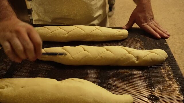 Professional baker's hands using a lame to score traditional patterns onto raw baguettes before placing them in the oven for baking