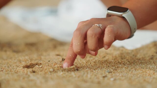Closeup hand tracing pattern in sand with smartwatch visible, traveler doodles playful grooves and explores texture with fingertip, sandy ring and nails catch sunlight, calm curious seaside moment