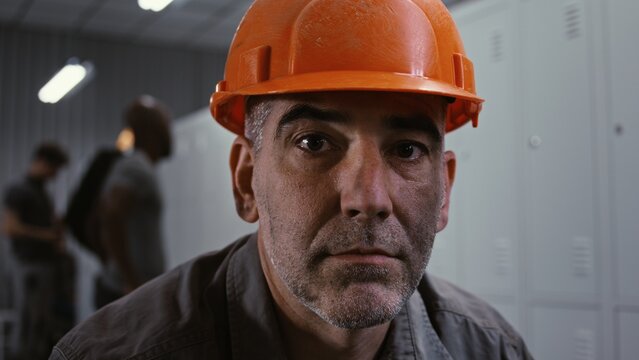 Close Up of Seasoned Factory Worker in Grey Work Jacket and Orange Helmet, Sitting on Bench and Looking Into Frame With Metal Lockers and Colleagues in Background. Industrial Preparation. Portrait.