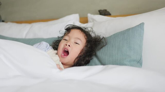 A sleepy young child yawns widely while waking up in a comfortable white bed, a candid moment of childhood fatigue representing rest and domestic wellbeing.