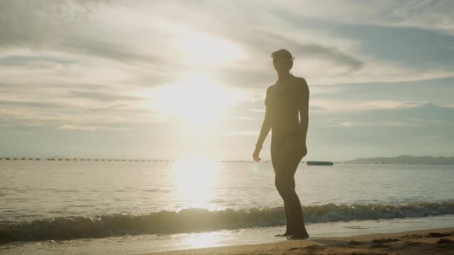 Silhouette walking on beach at sunset, pausing to gaze horizon, adjusting hair against golden light, calm waves lapping shore, contemplative traveler embracing solitude and reflection