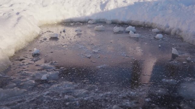 A dark frozen ice patch is surrounded by white snowbanks and scattered with small chunks of hard snow, creating a close winter view of rough icy texture outdoors. High quality 4k footage