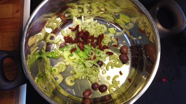 Stovetop close-up of sliced celery, sun-dried tomatoes and olives in a stainless cooking pot with olive oil, capturing fresh Mediterranean ingredients being prepared for a savory recipe.