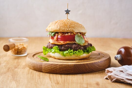 A ready-made burger with a beef patty, melted cheese, crispy onions, tomato, and lettuce on a wooden board against a wooden background.