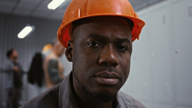 Close Up of African American Male Worker in Orange Safety Helmet, Staring With Serious and Focused Look Directly Into Lens Inside Factory Locker Room. Powerful Gaze and Authentic Expression. Portrait.