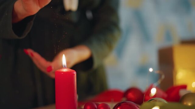 Hands protecting candle while wrapping present, careful gift wrapper shielding flame from draft, polished nails and delicate gesture, red candle glow, ornaments and lights on tabletop, intimate