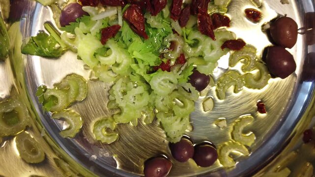 Fresh sliced celery tossed with Kalamata olives, chopped sun-dried tomatoes and olive oil on a reflective stainless steel tray, evoking rustic Mediterranean flavors, bright greens, and simple healthy 