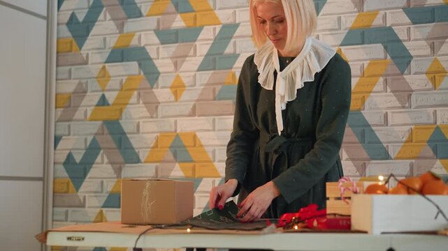 Caucasian woman wrapping christmas present, artisan shop owner carefully ties ribbon around kraft box on patterned brick wall, warm string lights glow, scissors and tape on wooden table, red bows