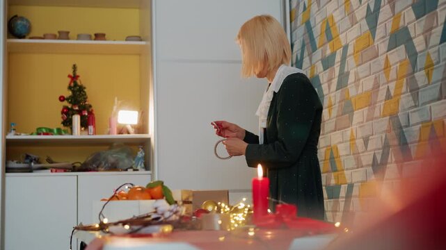 White woman preparing christmas present at cozy kitchen table with small decorated tree, glowing candle, fairy lights, mosaic wall, arranging ribbon and paper, cutting tape with scissors, festive