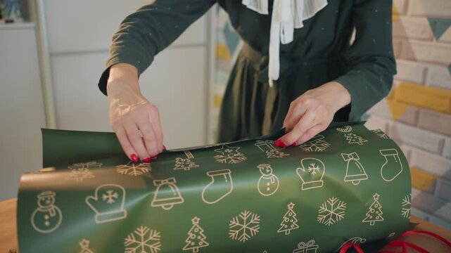 Hands wrapping green christmas present on wooden table, festive gold icons on paper, red nail polish, adhesive tape and scissors visible, folding precise edges, closeup of female crafter preparing