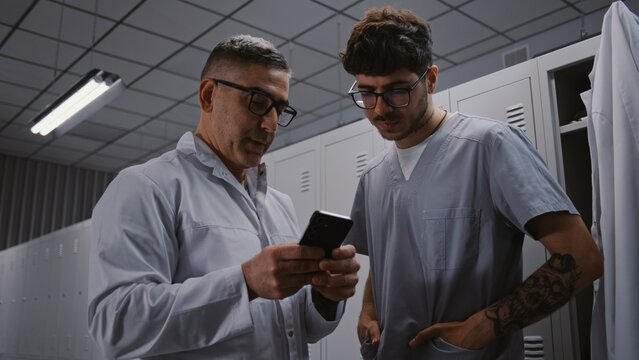 Senior Surgeon Mentors Younger Intern With Tattoos, Using Smartphone to Review Interns Scientific Research Paper and Findings in Staff Changing Area. Medical Education and Mentorship Concept.