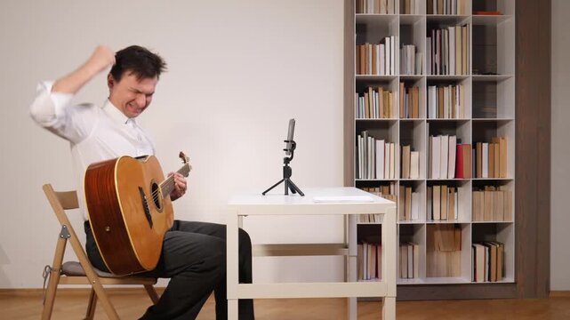 Man practicing acoustic guitar during an online remote lesson, watching the screen of a mobile phone placed on a portable tripod on a white desk, with a bookshelf visible in the background