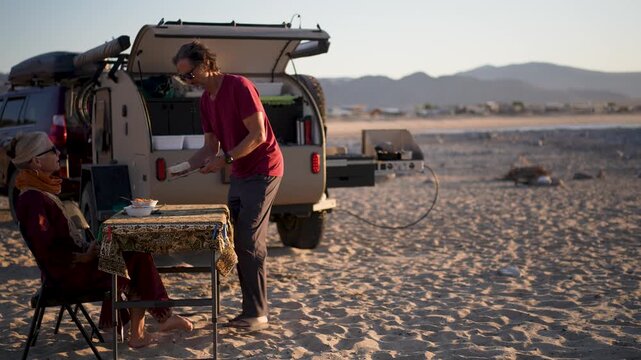 Mature man and woman share a meal at a table outside their teardrop camper trailer while enjoying the sunset on a beach.