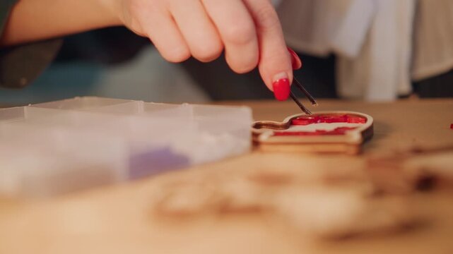 Closeup hands decorating Christmas ornament, precise tweezers placement, bead organizer, wooden cutouts, red lacquer nails, warm studio light, careful gift finishing, holiday craft detail,