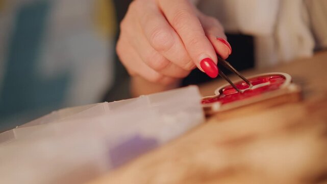 Closeup hands painting Christmas ornament, tweezers. Female crafter with red manicure uses tweezers to place tiny embellishments on round shiny ornament on wooden table, soft warm bokeh background,