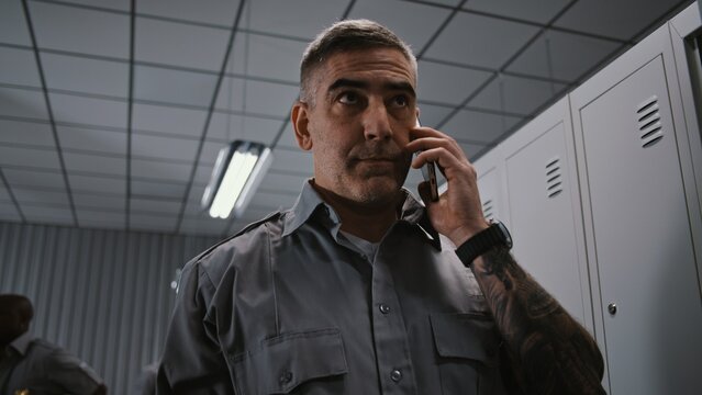Security Officer With Prominent Arm Tattoos Stands by Grey Metal Locker, Holding Smartphone and Having Serious Conversation After Workday Shift. Workplace Communication Concept in Staff Changing Room.