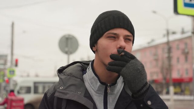 Pensive youth pauses with cigarette outdoors. Urban scene of young man awaiting bus on chilly day. Teenager stands at transit shelter under overcast sky with contemplative expression