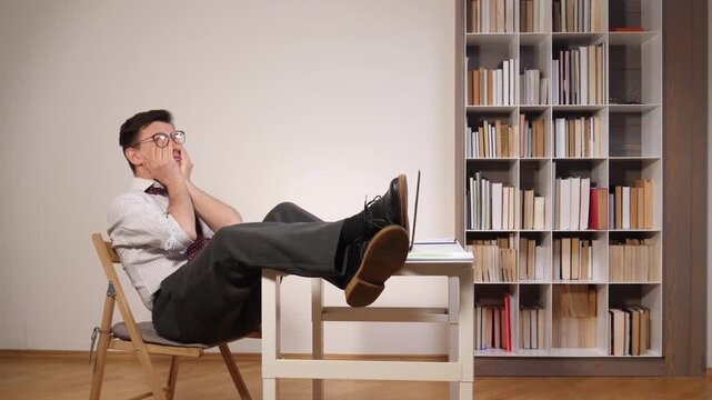 Tired businessman feeling burned out and frustrated, sitting with feet on a small desk beside a laptop in an office library, taking a weary break from work and deadlines
