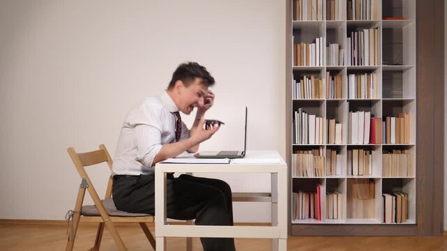 Businessman expressing anger and frustration while having a virtual conflict or dealing with a technical problem at his desk in an office environment with a bookshelf