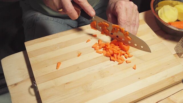 Close-up of a person's hands dicing fresh carrots with a sharp knife on a wooden board.