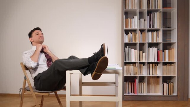 Young businessman taking a break from work, leaning back in a chair with his feet resting on a desk, looking upwards and contemplating in an office environment