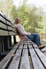 Fototapeta premium Selective focus view of a wooden park bench with a blurred caucasian woman resting in the background, ideal for solitude, reflection and mental wellness concepts.
