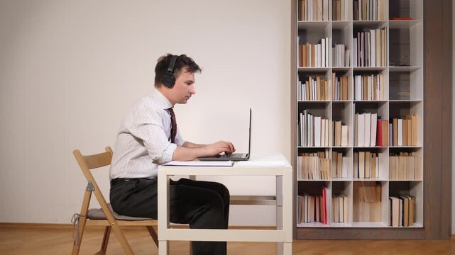 Professional man wearing headphones and concentrating while working remotely on a laptop at a desk in a home office with a bookshelf in the background