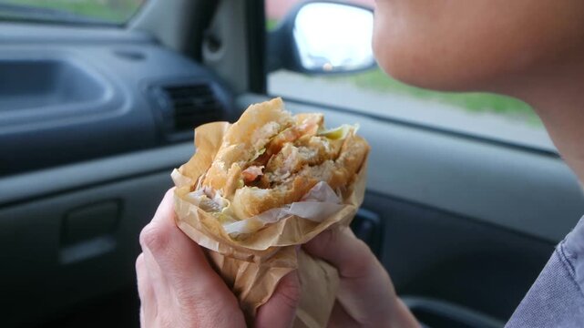 Close up person sitting inside a car and eating a large, greasy hamburger wrapped in brown paper, a quick and unhealthy meal for a busy lifestyle representing a common fast food habit