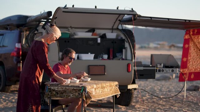Couple enjoys a meal while camping by the beach in Baja California during sunset next to their teardrop camper trailer.
