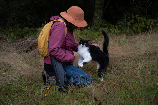 Una mujer con sombrero y mochila acaricia a un gato de color blanco y negro en medio del campo.
