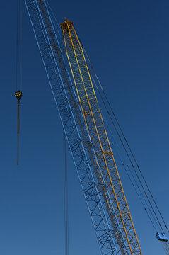 A tall construction crane with a lattice boom and hanging hook rises against a clear blue sky