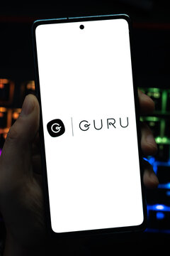 Punta Arenas, Chile &ndash; May 04, 2026: Close-up of a mobile device screen showing the Guru enterprise search and knowledge management platform brand, held by a user in a dark room with vibrant keyboard