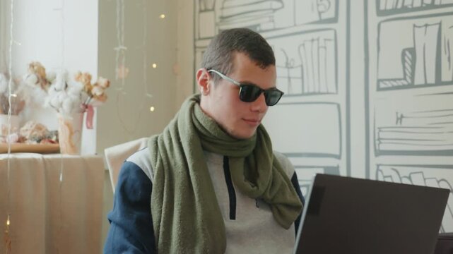 Caucasian man wearing sunglasses typing at laptop in bright windowed studio, green scarf and casual jacket, working as freelance designer and startup founder, mood reflective and stylish, soft natural